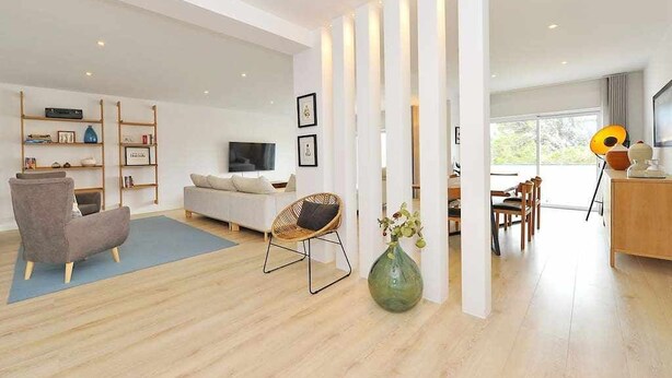A modern living room with a sofa, armchair, and a dining area with chairs and a table. - Prestige Sunset Carcavelos Beach - Checkinhome (Carcavelos)