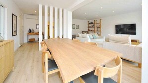 A spacious living area with a long wooden dining table, a white sofa, and a flat-screen TV mounted on the wall. - Prestige Sunset Carcavelos Beach - Checkinhome (Carcavelos)