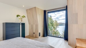 Ein Schlafzimmer mit Bett, Kommode, Fenster mit Blick auf einen See und Bäume sowie Holzboden.