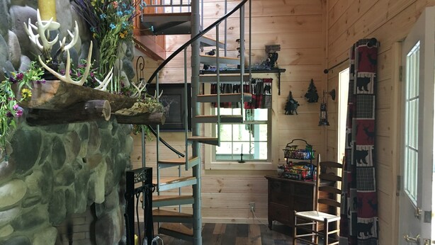 A rustic cabin interior with a spiral staircase, stone wall, wooden floors, and various decorations including antlers and a sign.