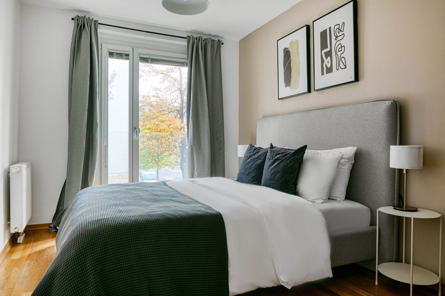 Ein modernes Schlafzimmer mit einem großen Bett, grauen Vorhängen, einem Fenster mit Blick auf Bäume und zwei gerahmten Kunstwerken an der Wand.