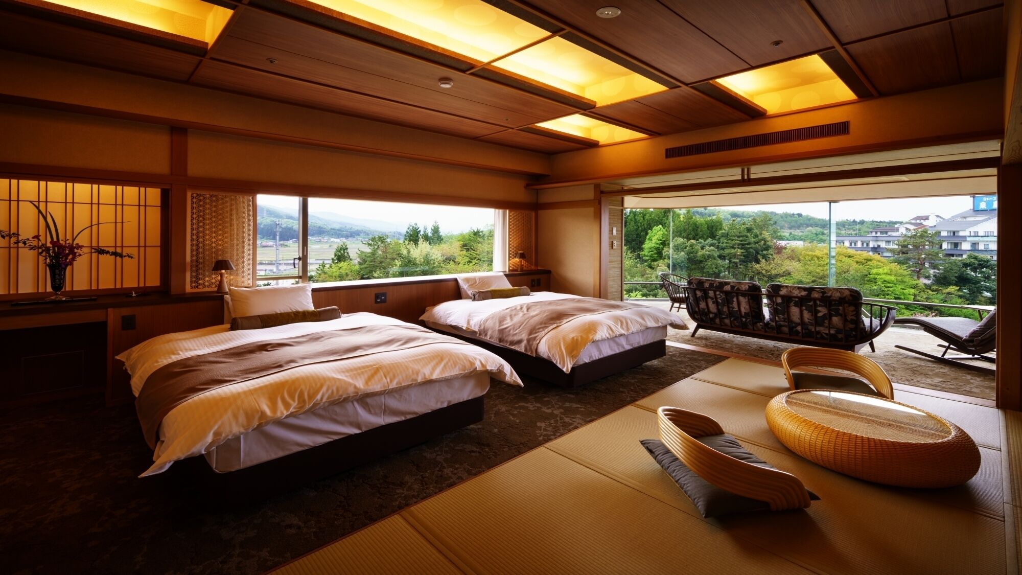 A hotel room with two beds, tatami mats, a balcony with chairs, and a view of greenery.
