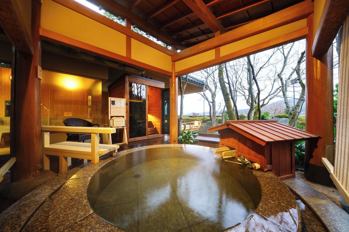 Un onsen japonés tradicional con arquitectura de madera, un baño de aguas termales y vistas a árboles y montañas.