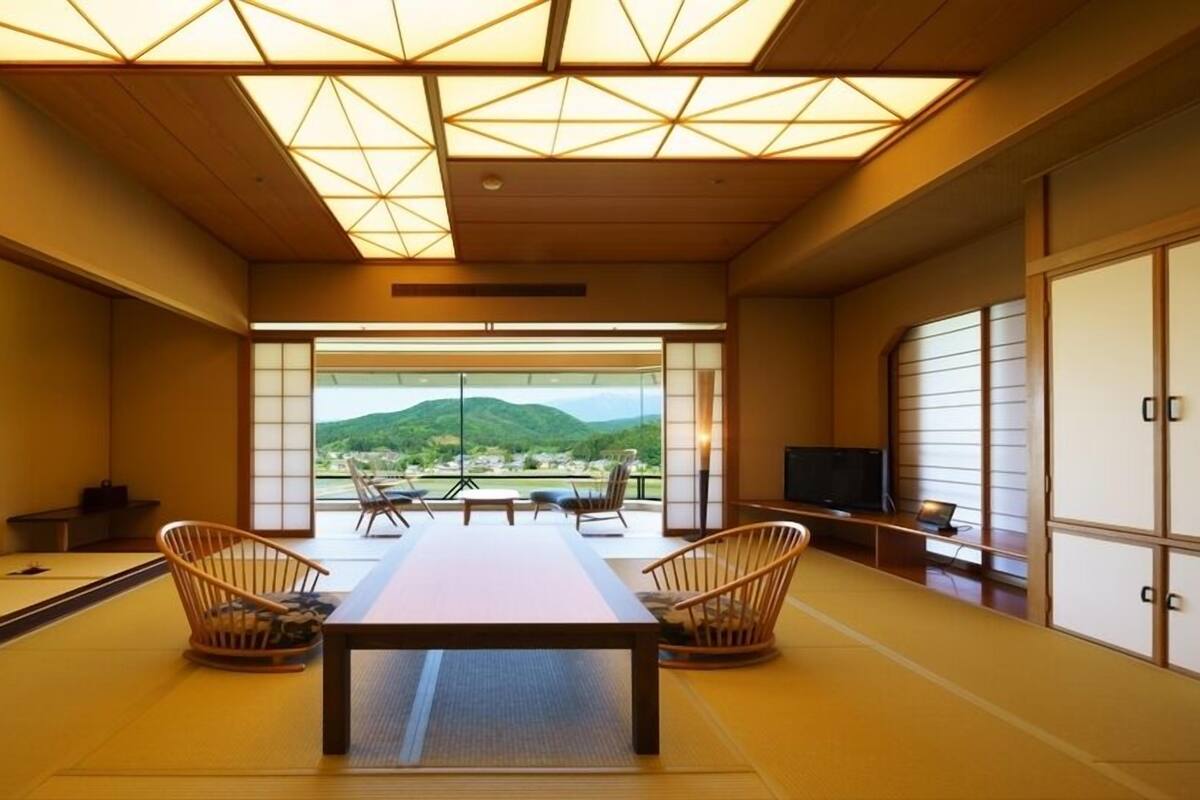 A traditional Japanese room with tatami flooring, a low table, two chairs, a TV, and a view of a mountain landscape.