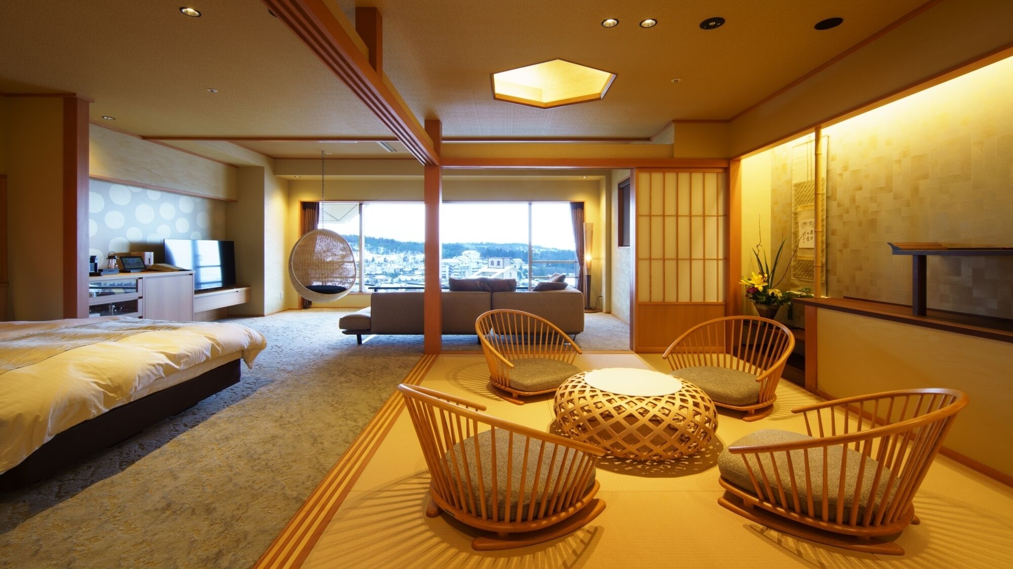 A hotel room with a bed, a sofa, a dining table with chairs, a hanging chair, and a view of the city.