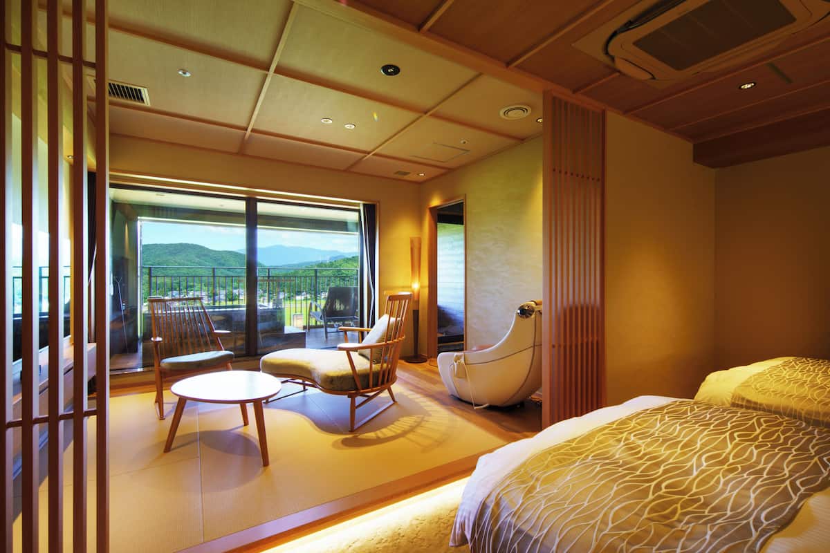 A hotel room with a bed, a chair, a small table, and a view of the outdoors.