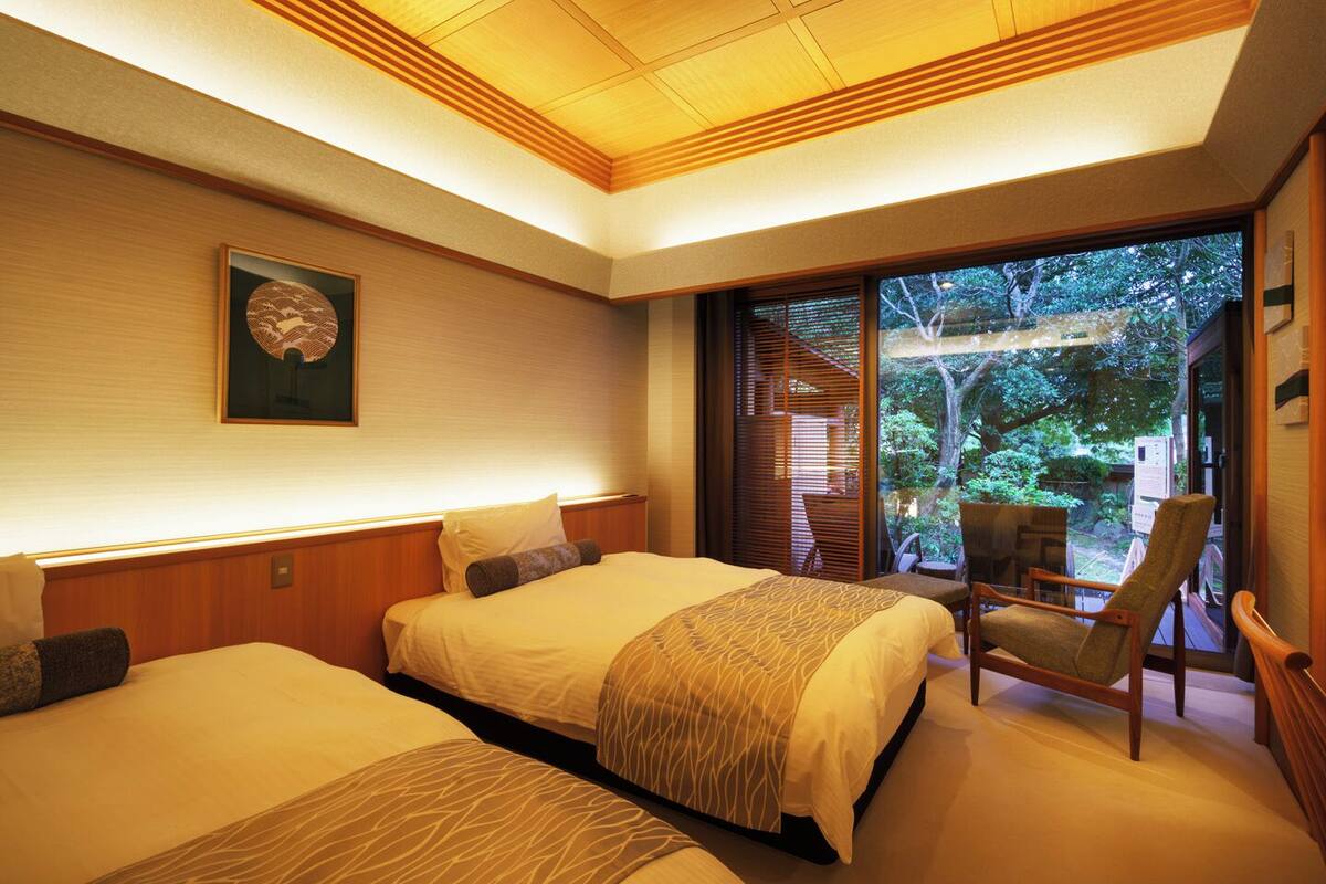A hotel room with two beds, a chair, a large window with a view of trees, and a ceiling with wooden paneling.