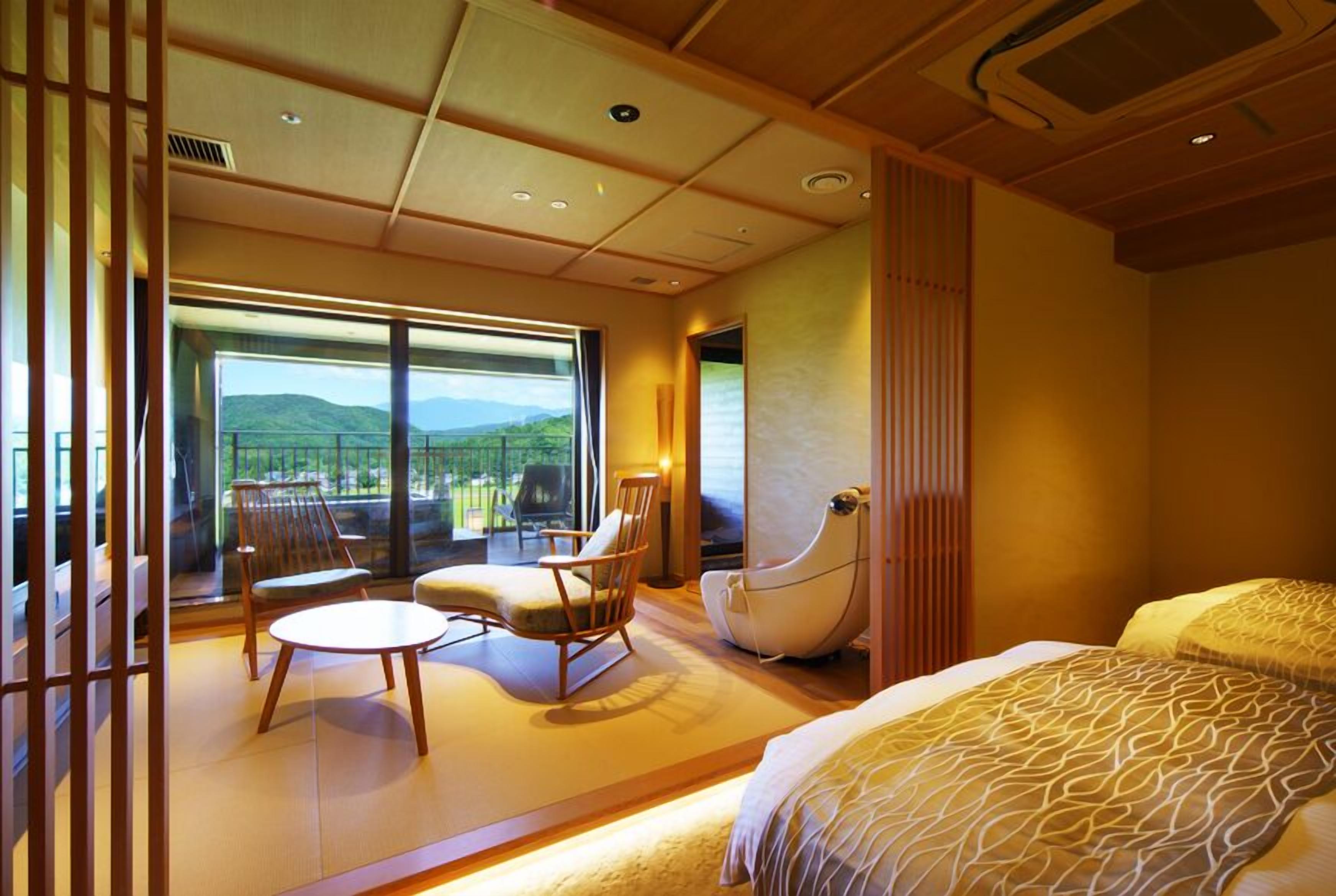 A hotel room with a large sliding glass door leading to a balcony, a bed with patterned bedding, a round table, and two chairs.