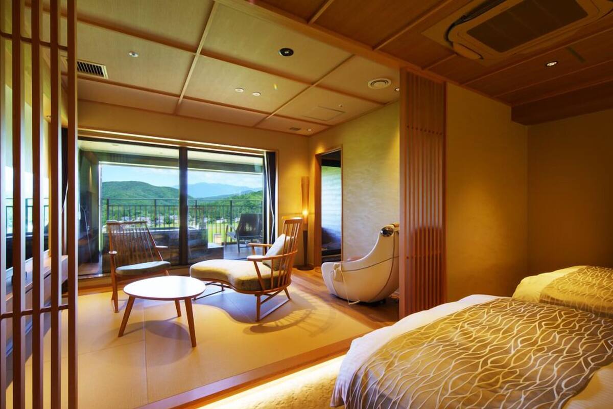 A hotel room with a large sliding glass door leading to a balcony, a bed with patterned bedding, a round table, and two chairs.