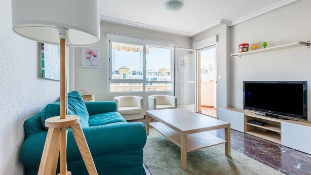Ein modernes Wohnzimmer mit einem türkisblauen Sofa, einem Holztisch, einem Fernseher auf einem Lowboard und einem großen Fenster mit Blick auf Gebäude.