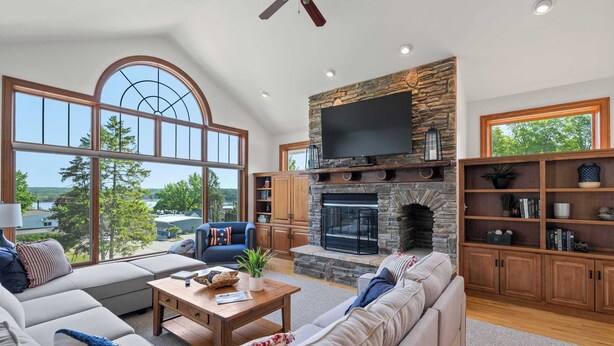 A spacious living room with a stone fireplace, large windows, and a television. - Walk to Shops! 3 Story Condo w/ Views + Elevator (Fish Creek)