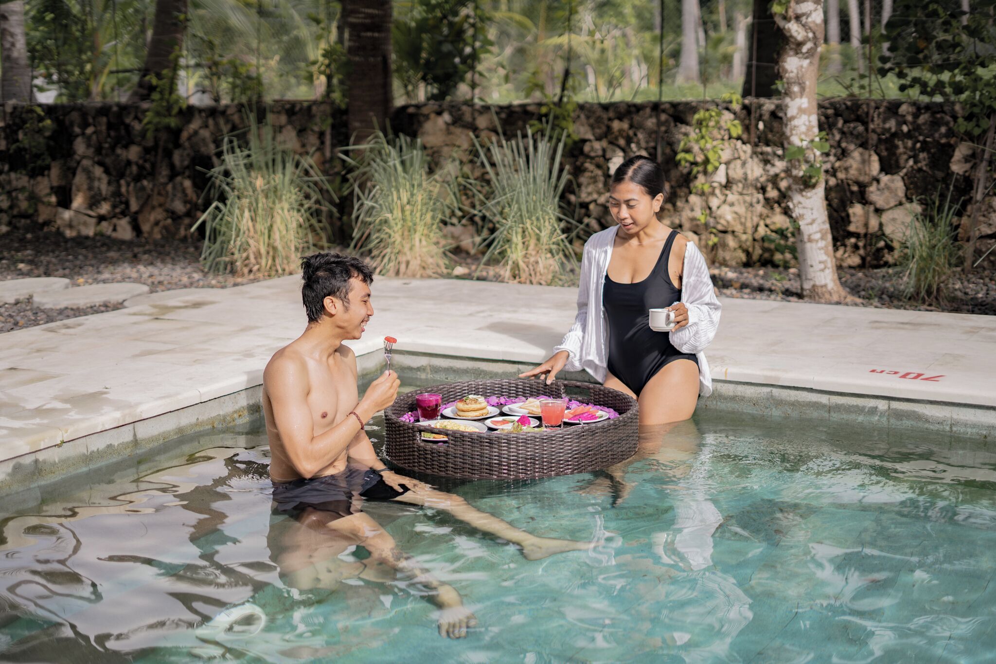 A man and a woman sitting by a pool with a basket of food.