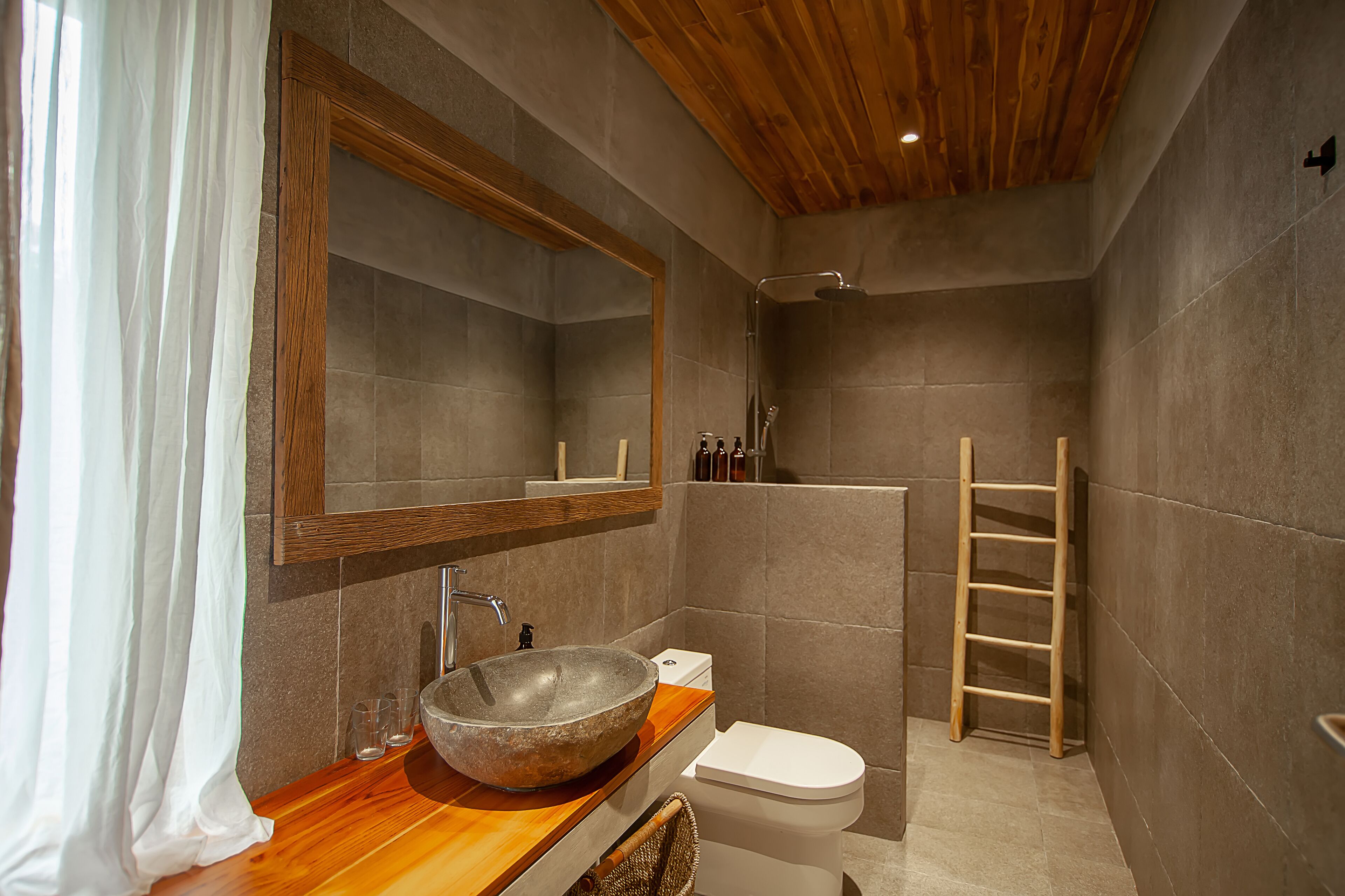 A modern bathroom with a wooden vanity, a large mirror, a stone sink, a toilet, and a shower area.
