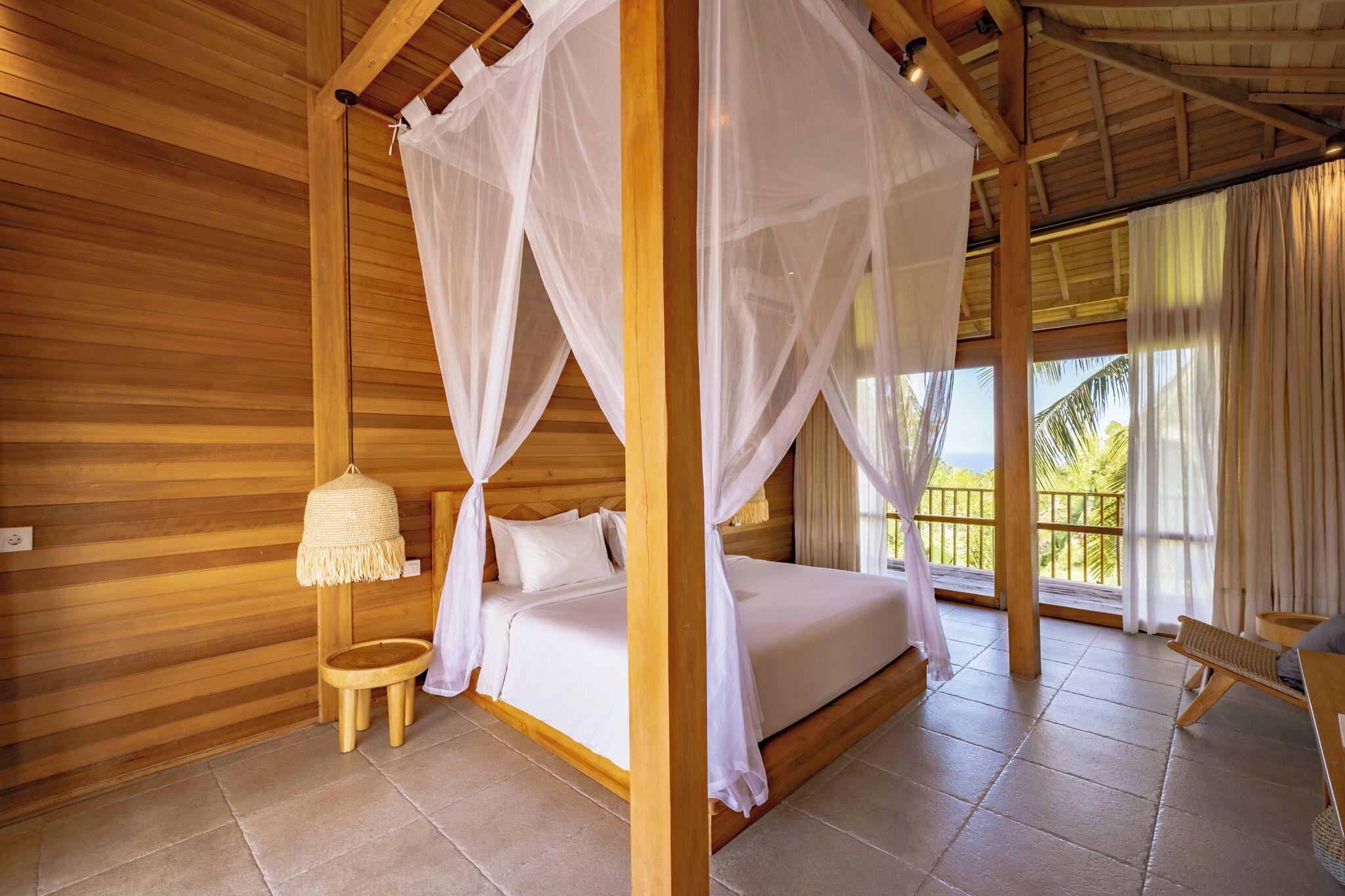 Ein Zimmer mit Holzboden, einem Bett unter einem Baldachin, einem kleinen Hocker und einem Korbstuhl.