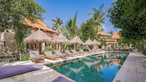 A pool area with lounge chairs and thatched umbrellas, surrounded by palm trees and a building with a red roof. - Amrani Diamond Beach Villas & Pool (Penida Island)
