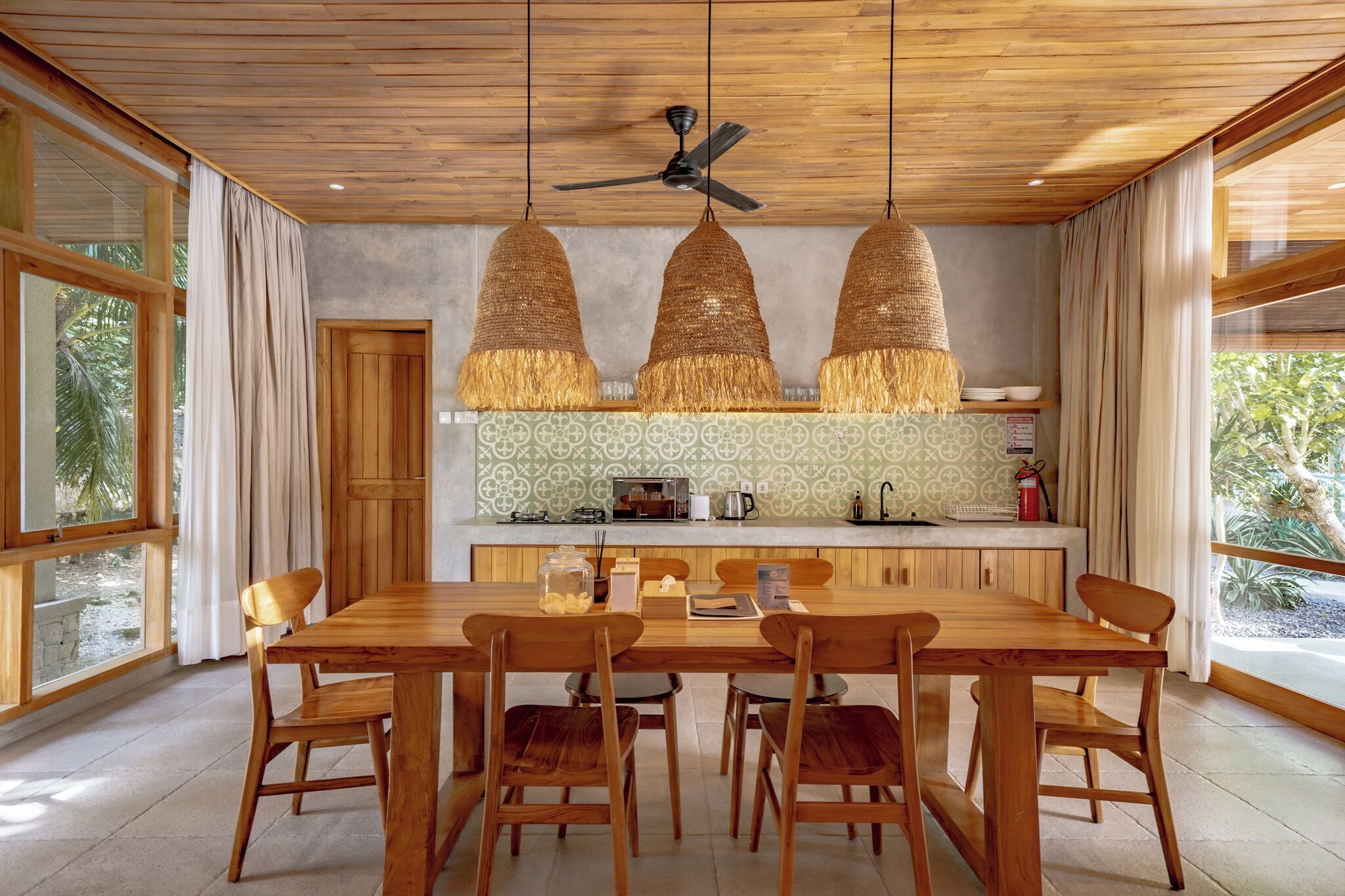A wooden dining table with chairs, a ceiling fan, and hanging woven light fixtures in a room with large windows and a kitchen area.
