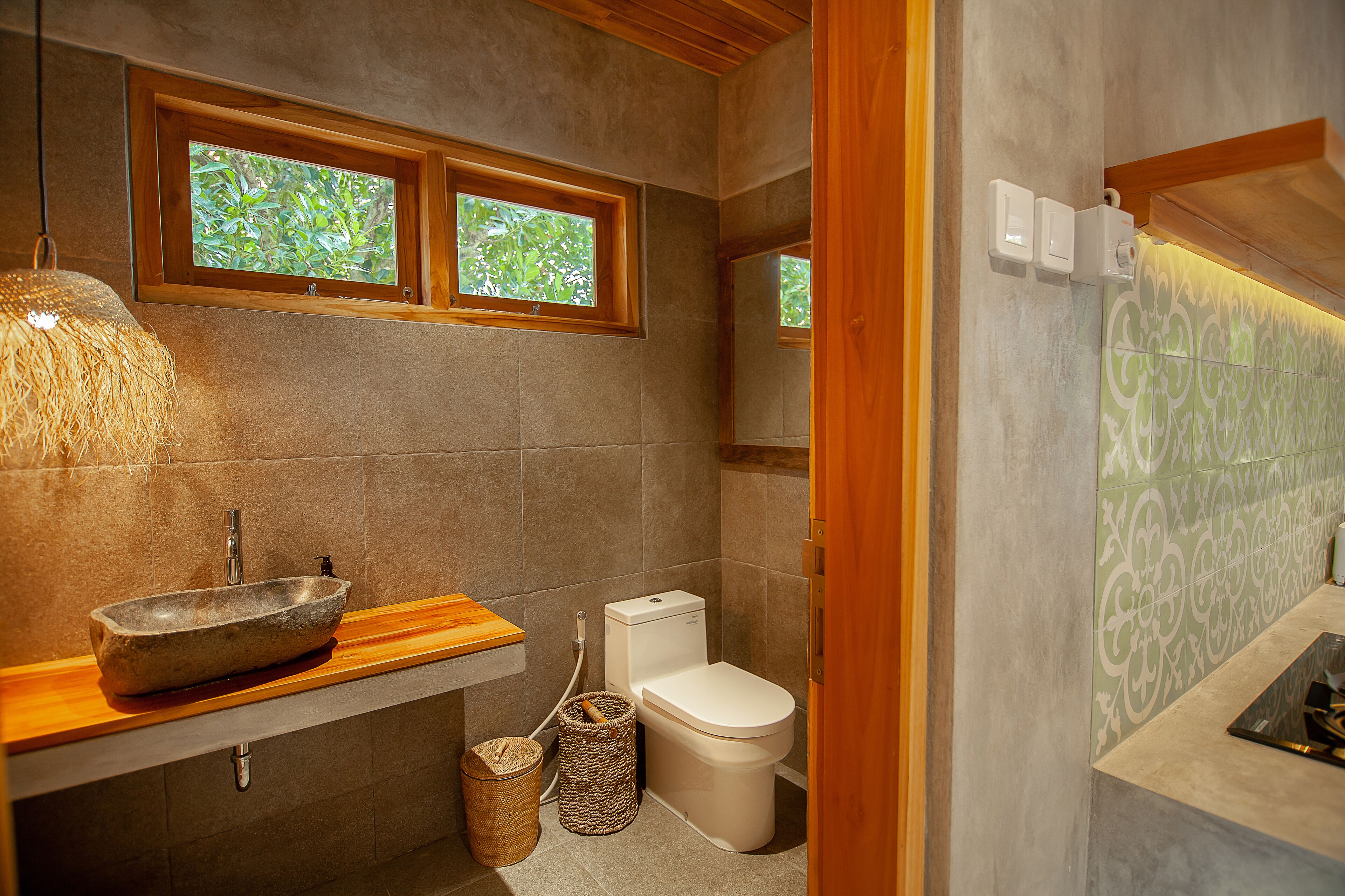 A bathroom with a stone sink, wooden countertop, and a toilet with a white tank. There are two windows above the sink and a towel rack on the wall.