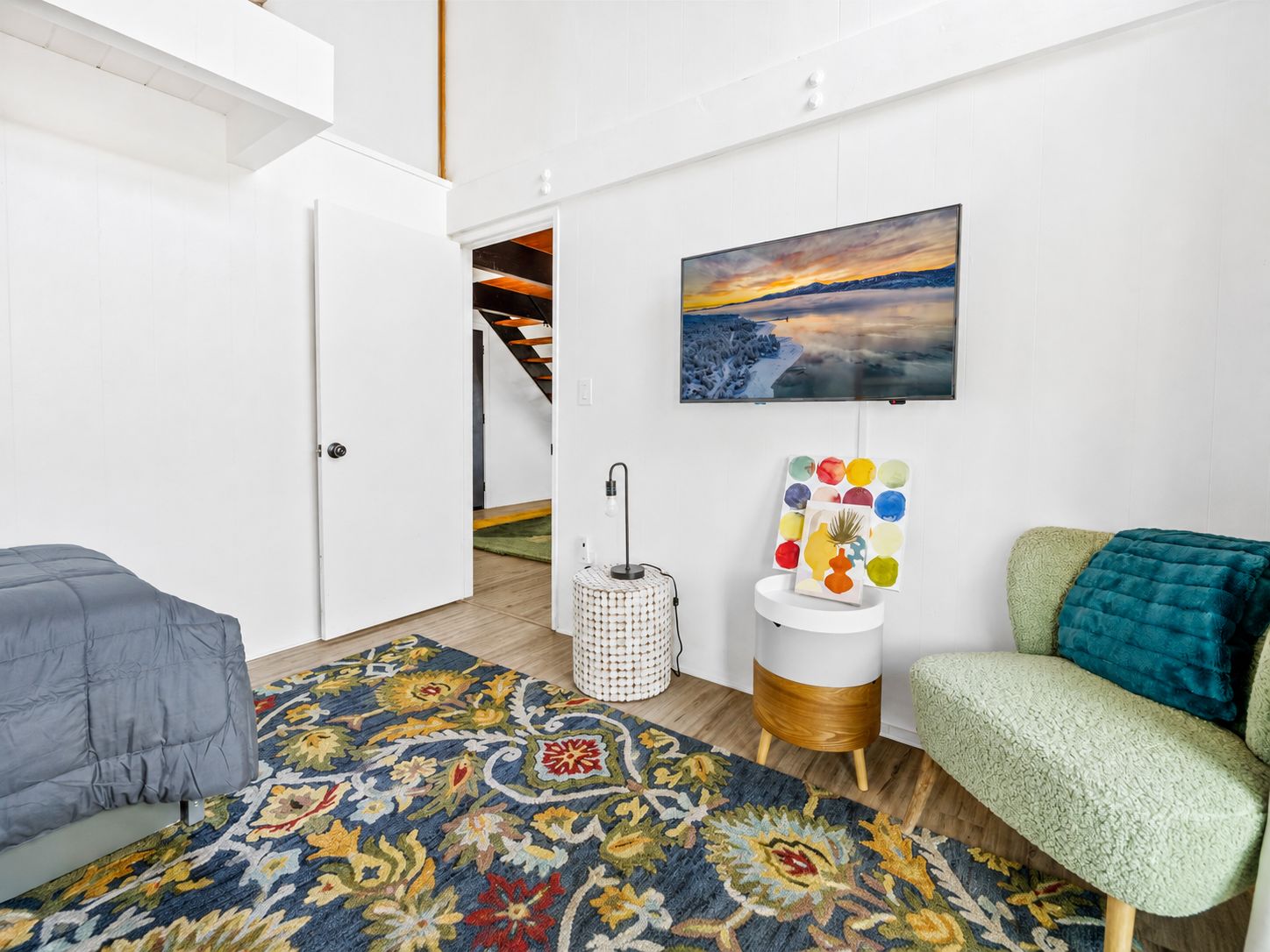 A modern living room with a patterned rug, a green sofa, a gray bed, and a painting of a sunset.