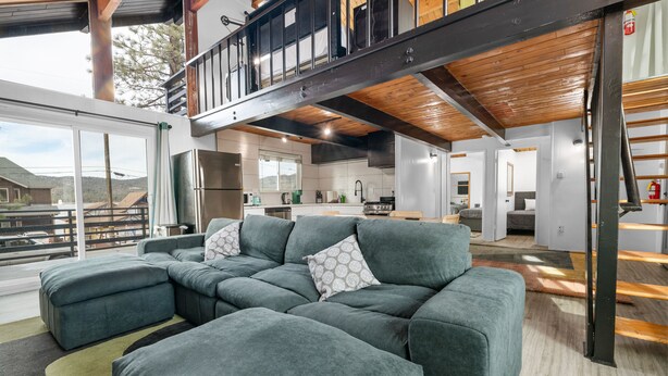 A modern living room with a sectional sofa, a balcony with a wooden railing, and a kitchen in the background.