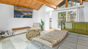 A spacious bedroom with a large bed, a wooden ceiling, a painting of a landscape, a cactus, and a view of the outside.