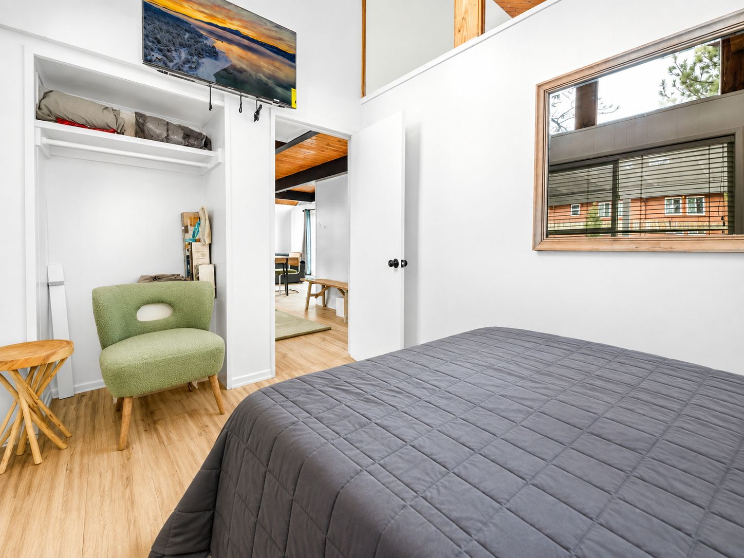 Une chambre avec un grand lit, un fauteuil vert, un plancher en bois, une armoire blanche et une fenêtre donnant sur des immeubles.