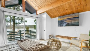 A bedroom with a large bed, a wooden ceiling, a view of the outdoors, a painting on the wall, and a bench.