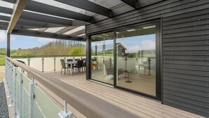 Ein überdachter Außenbereich mit Tisch und Stühlen, eine Glastür, die zu einer Terrasse führt, und ein Blick auf die umliegende Landschaft.