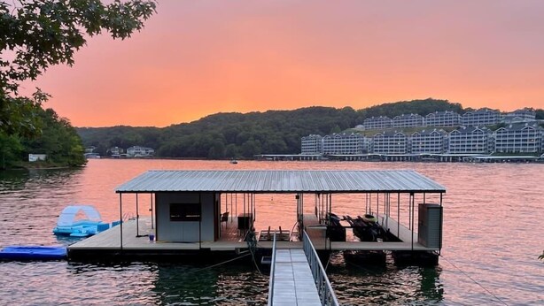 A docked houseboat with a covered deck and seating area, connected to a dock by a walkway.