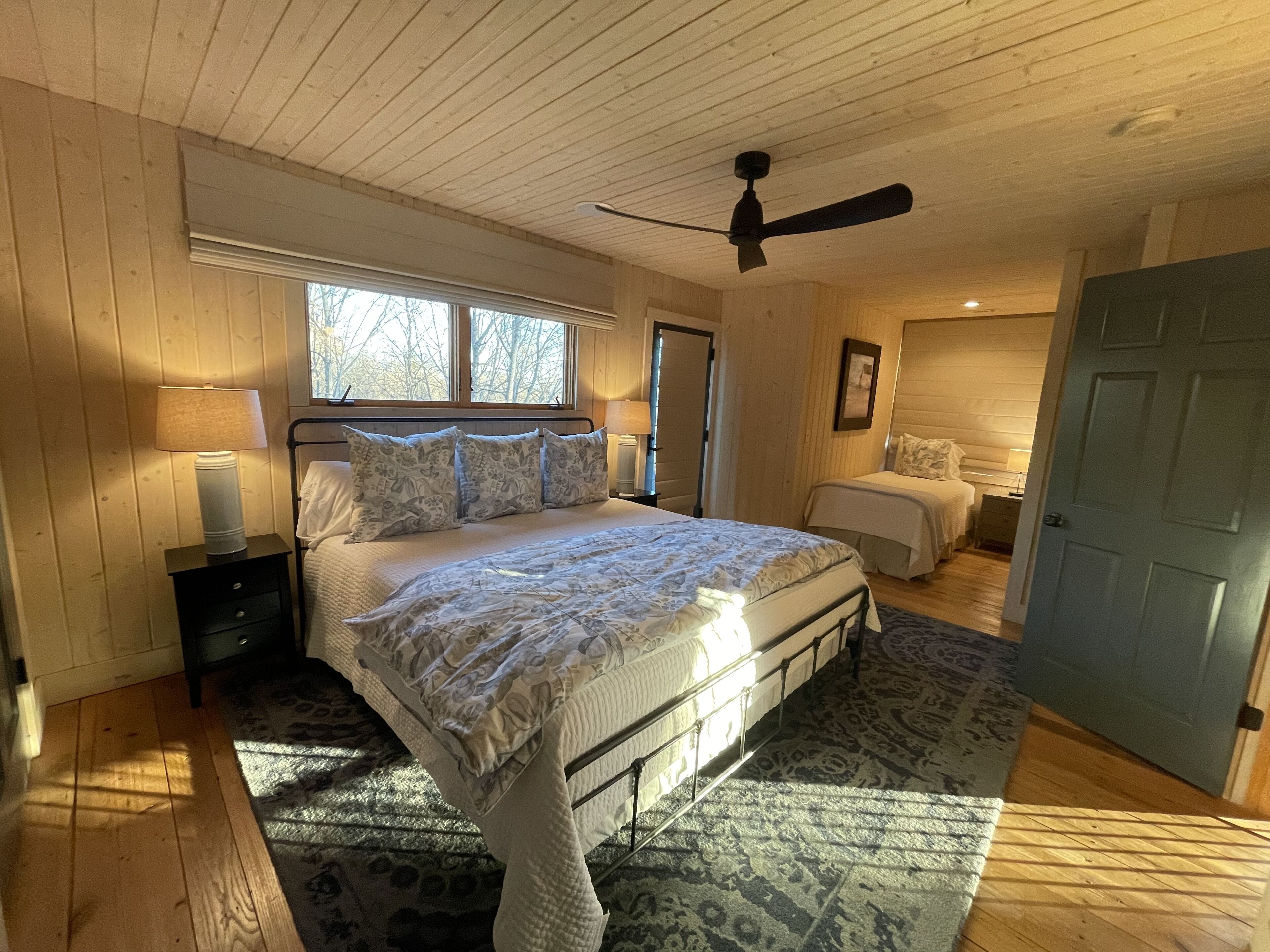 A bedroom with a large bed, a bedside table, a nightstand, a ceiling fan, a window, and a door.