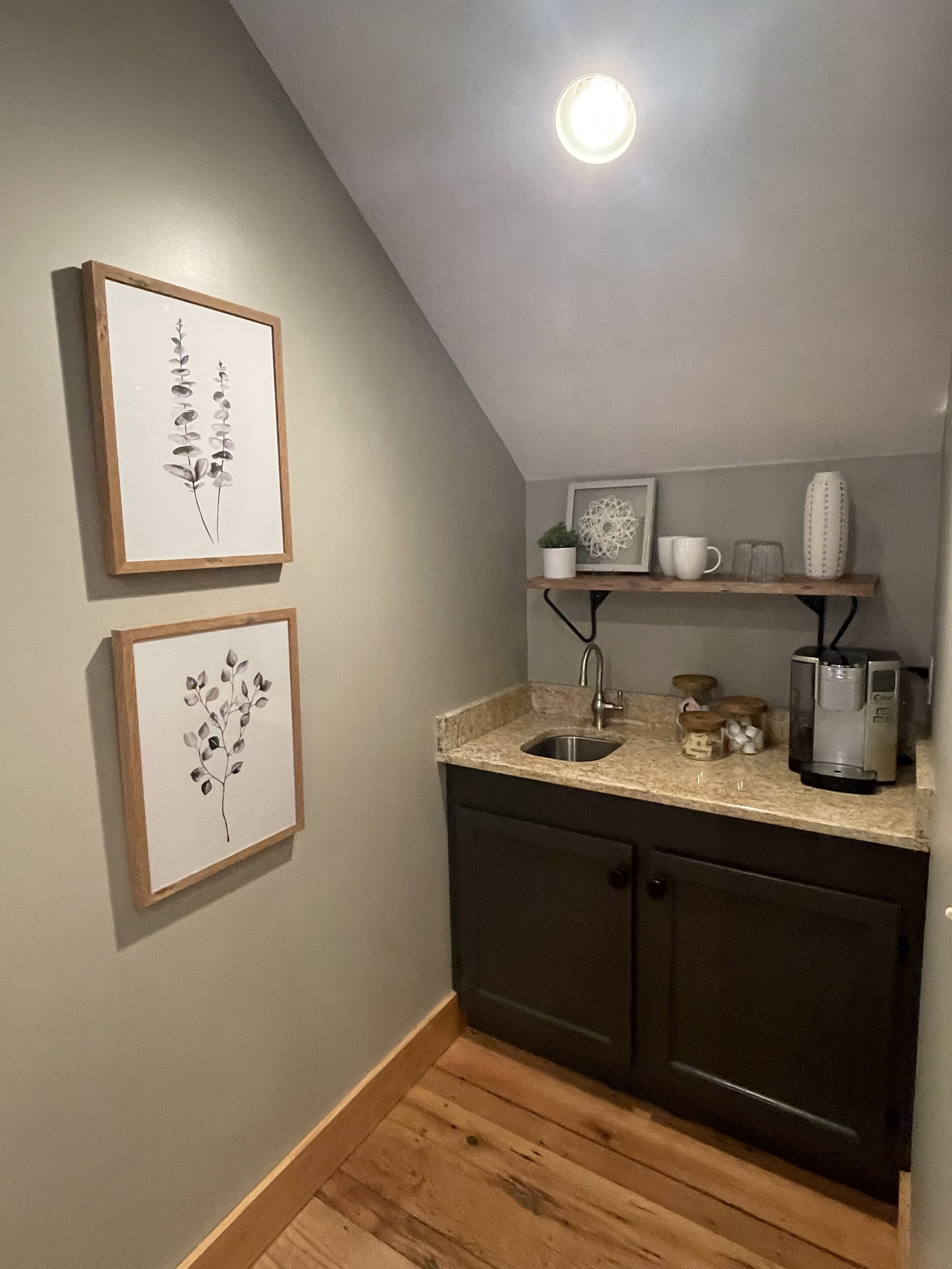 A small kitchen with a wooden countertop, a sink, and a coffee maker. There are two framed pictures on the wall and a shelf with a plant, a mug, and a vase.