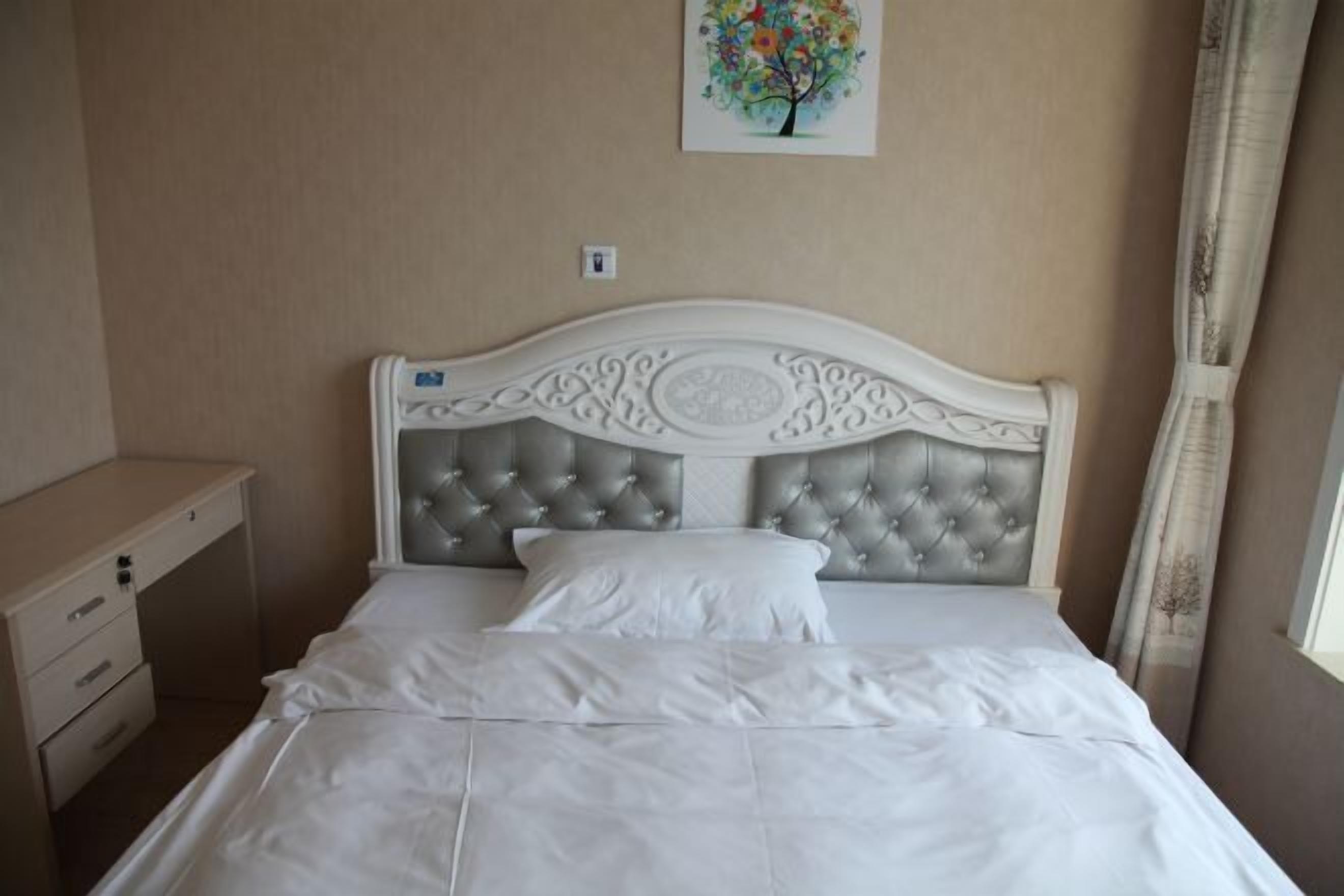 A neatly made bed with a tufted headboard, a bedside table with drawers, and a wall-mounted light switch.