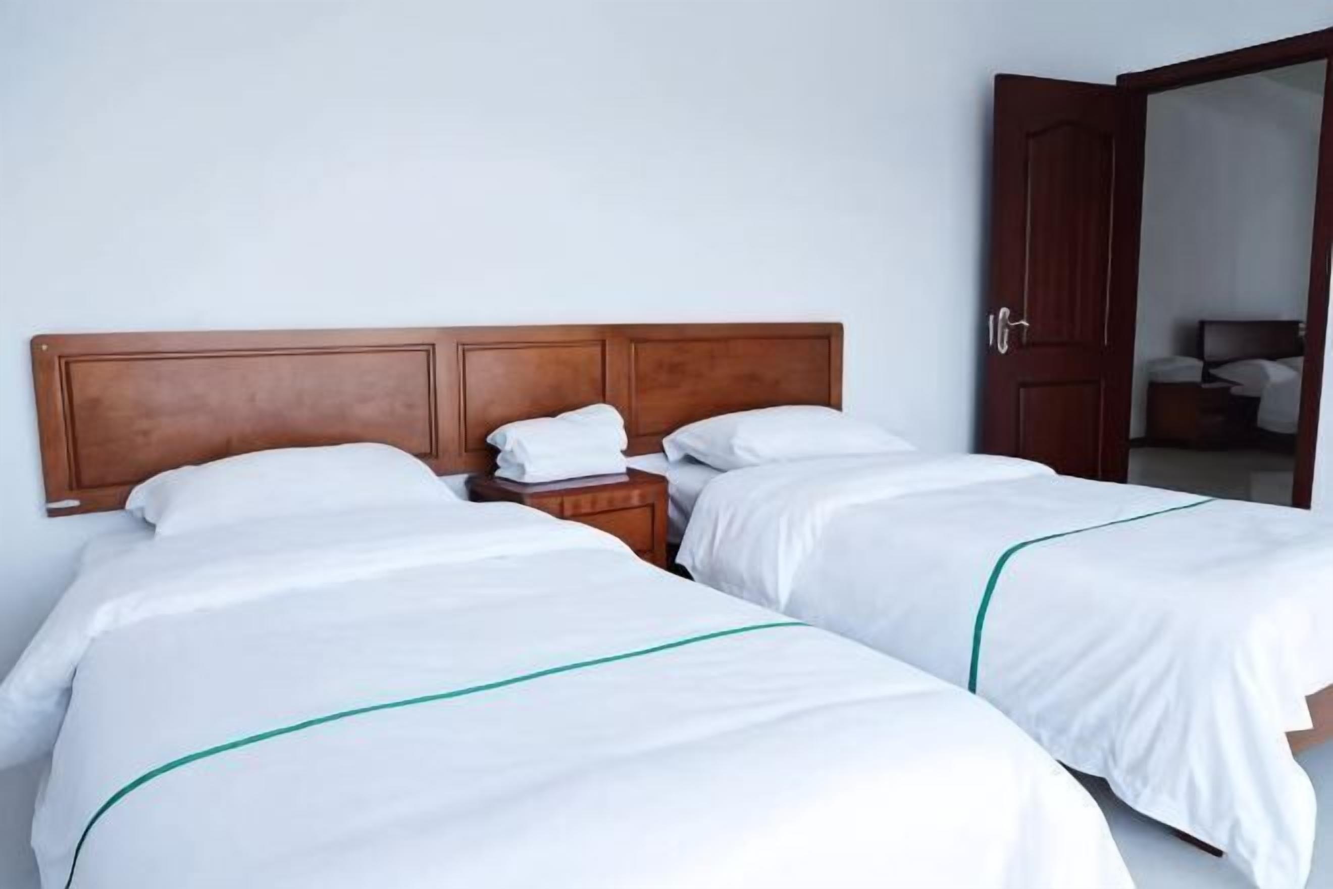 Quarto de hotel com duas camas, cabeceira de madeira, mesa de cabeceira e uma porta que dá para outro cômodo.