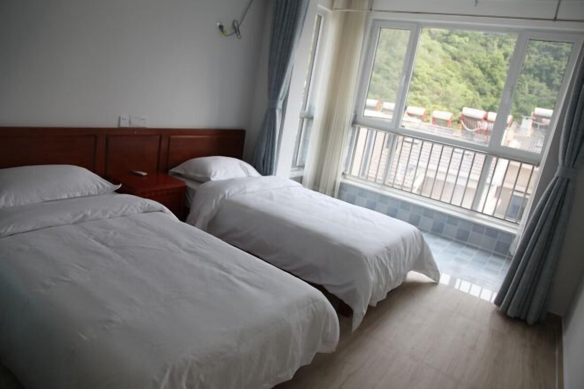 Habitación de hotel con dos camas, cabecera de madera, mesita de noche y un ventanal con vistas a zonas verdes y un balcón.