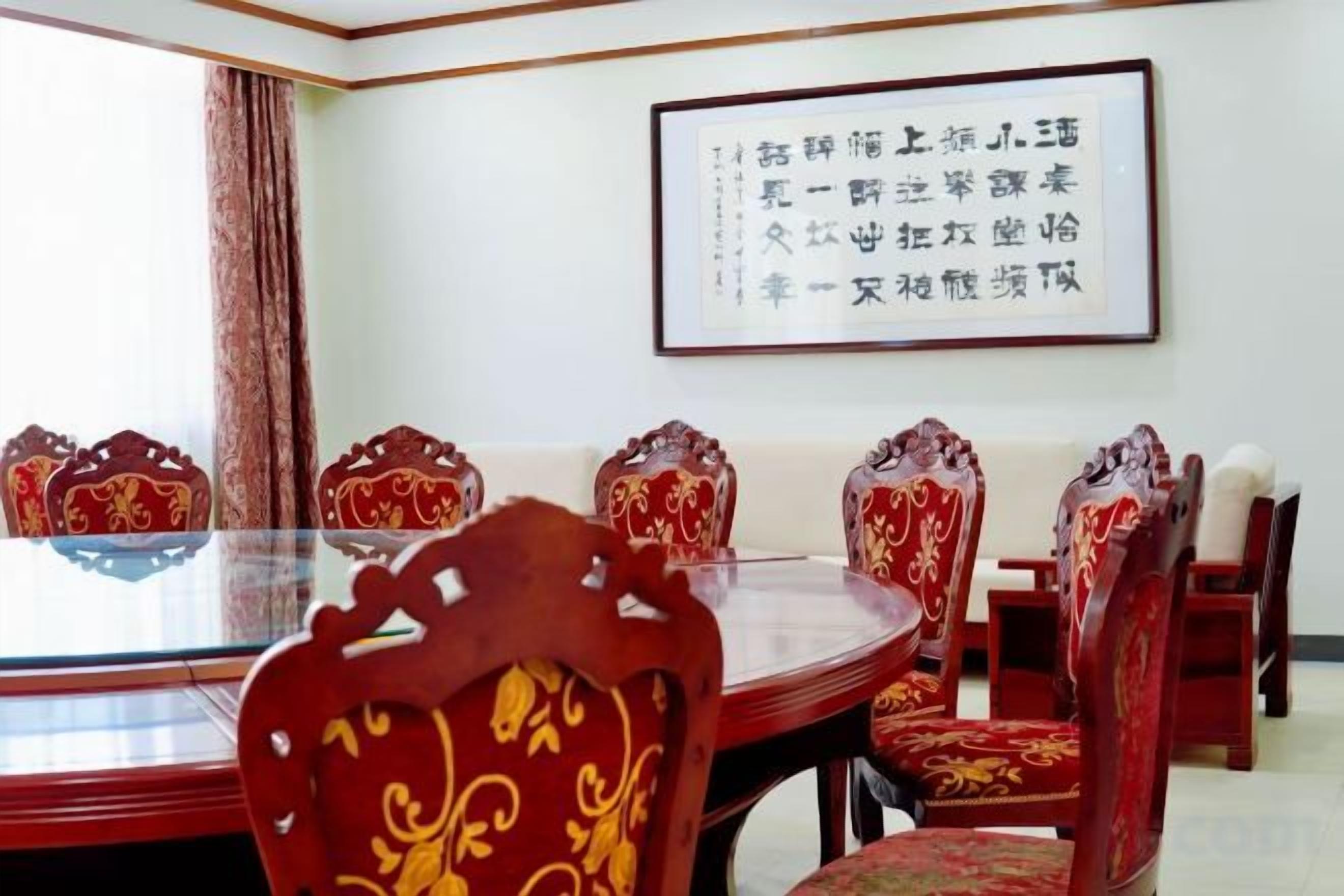 A conference room with a round glass table surrounded by ornate chairs, a framed calligraphy piece on the wall, and a window with curtains.