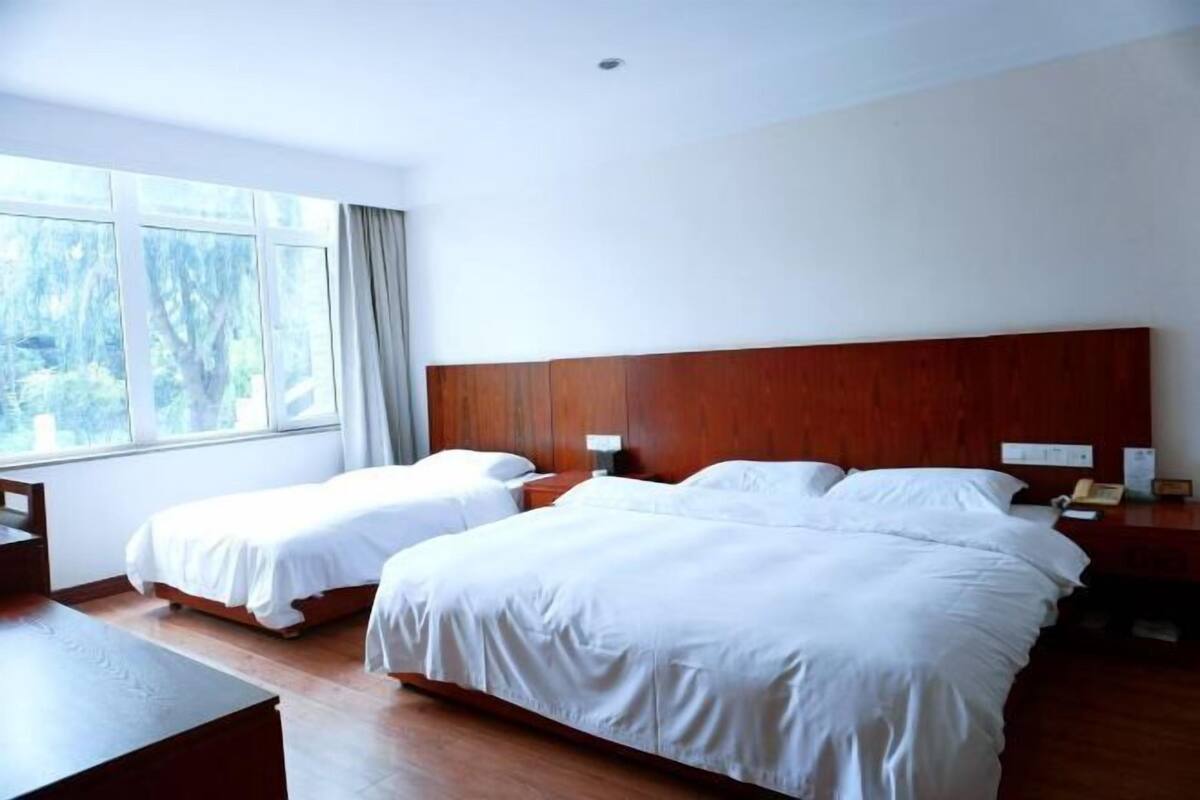 Een hotelkamer met twee bedden, een houten hoofdeinde, een nachtkastje, een bureau en een raam met uitzicht op bomen.