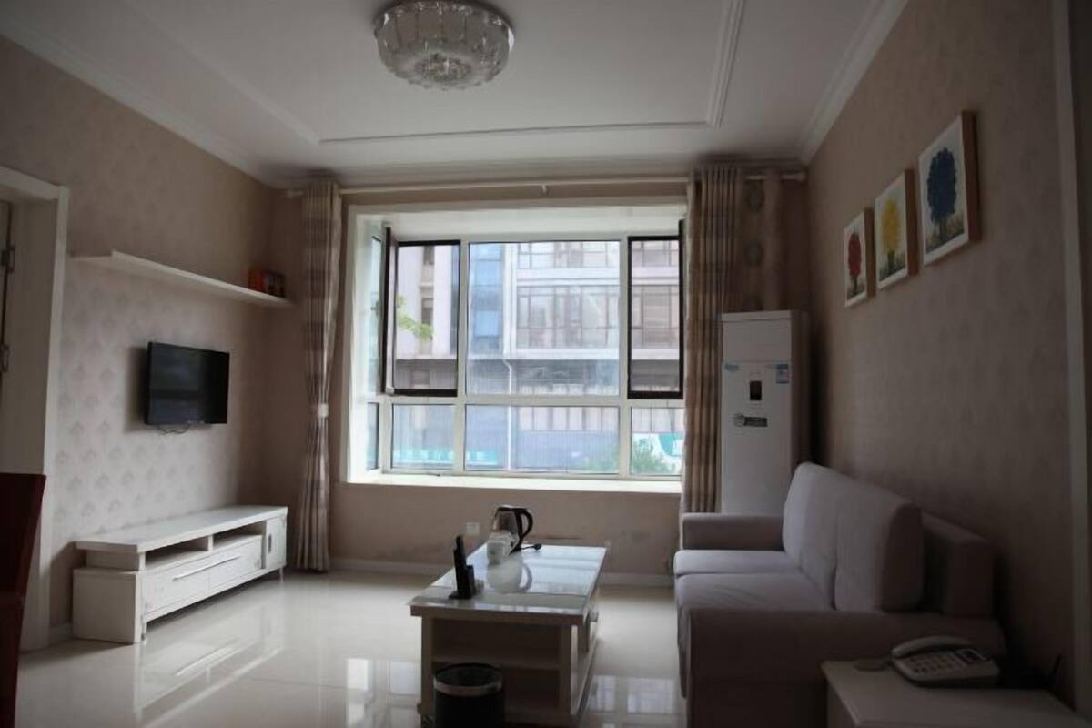 Een moderne woonkamer met een flatscreen televisie, een witte bank, een salontafel en een groot raam met gordijnen.