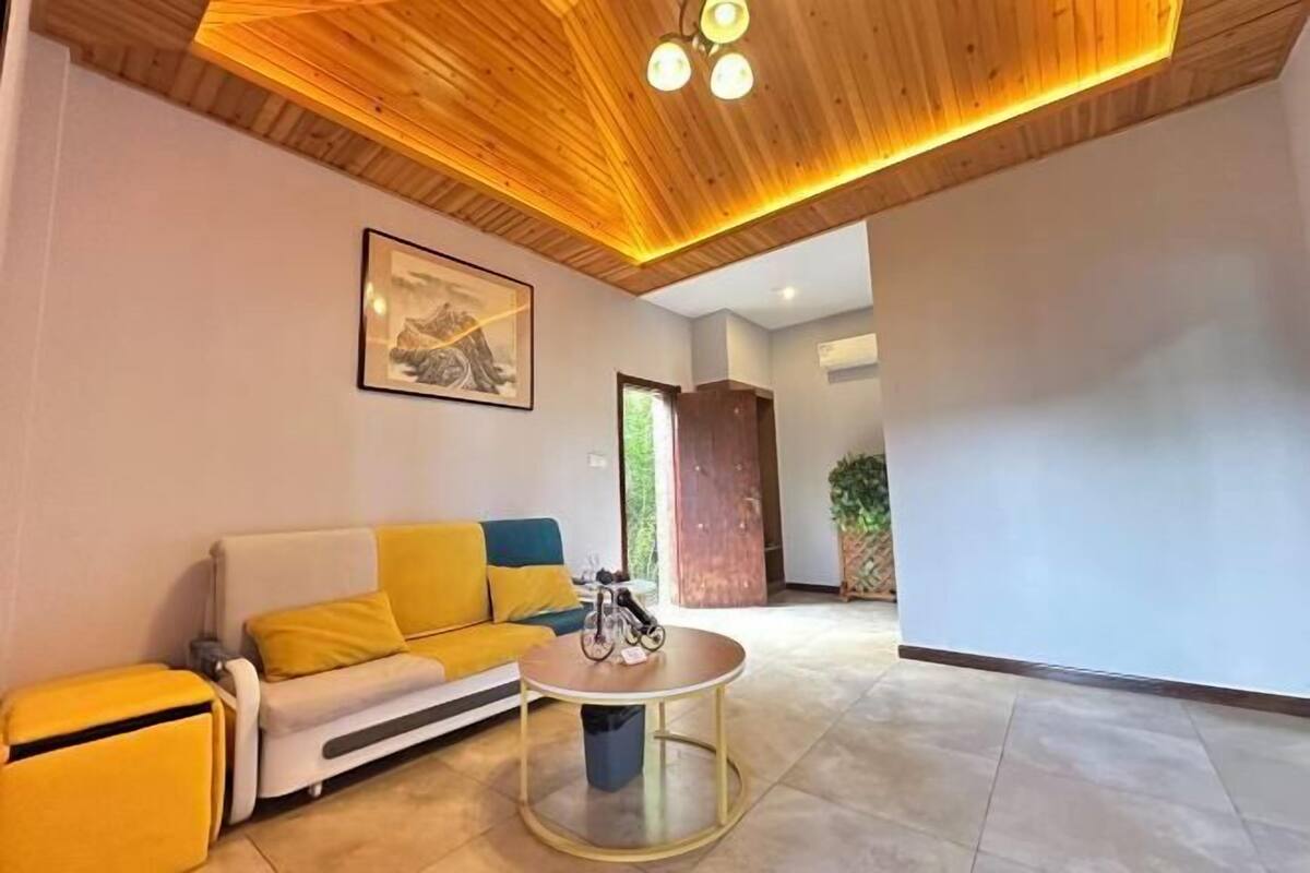 Een moderne woonkamer met een houten plafond, een bank met gele kussens, een ronde salontafel en een ingelijst schilderij aan de muur.