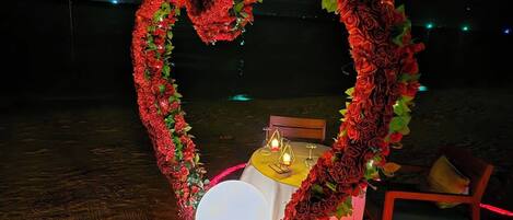 Un arreglo floral con forma de corazón sobre una mesa en la playa, con velas y un cuenco blanco, rodeado de pétalos de rosa.