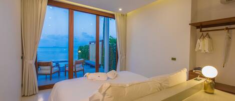 A hotel room with a large bed, a bedside table with a lamp, a view of the sea, and a balcony with chairs and a table.