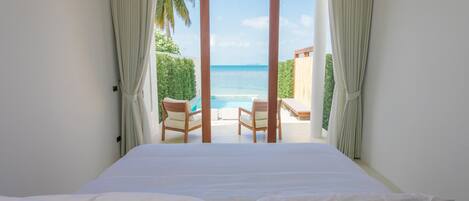 A bedroom with a large bed, a view of a pool and beach, and curtains on the windows.