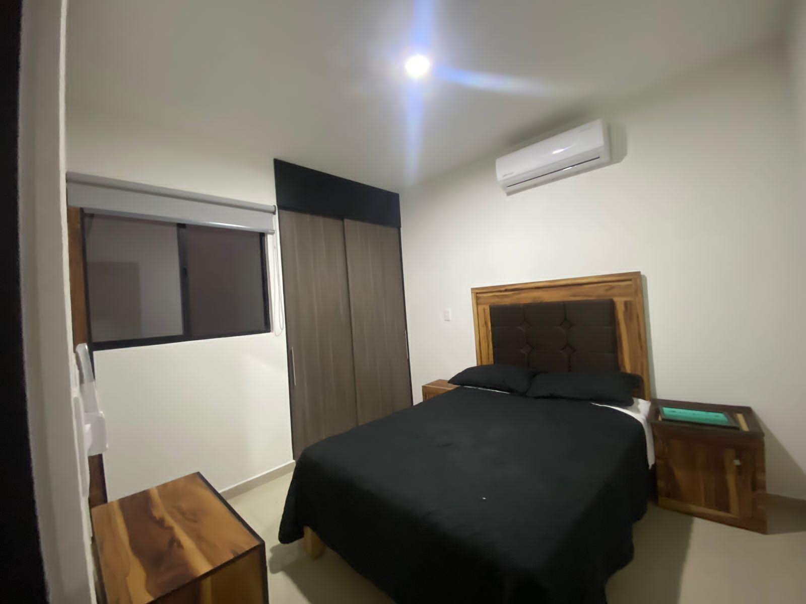 Un dormitorio con una cama, una mesita de noche de madera, una pequeña mesa de madera, una ventana y un equipo de aire acondicionado.
