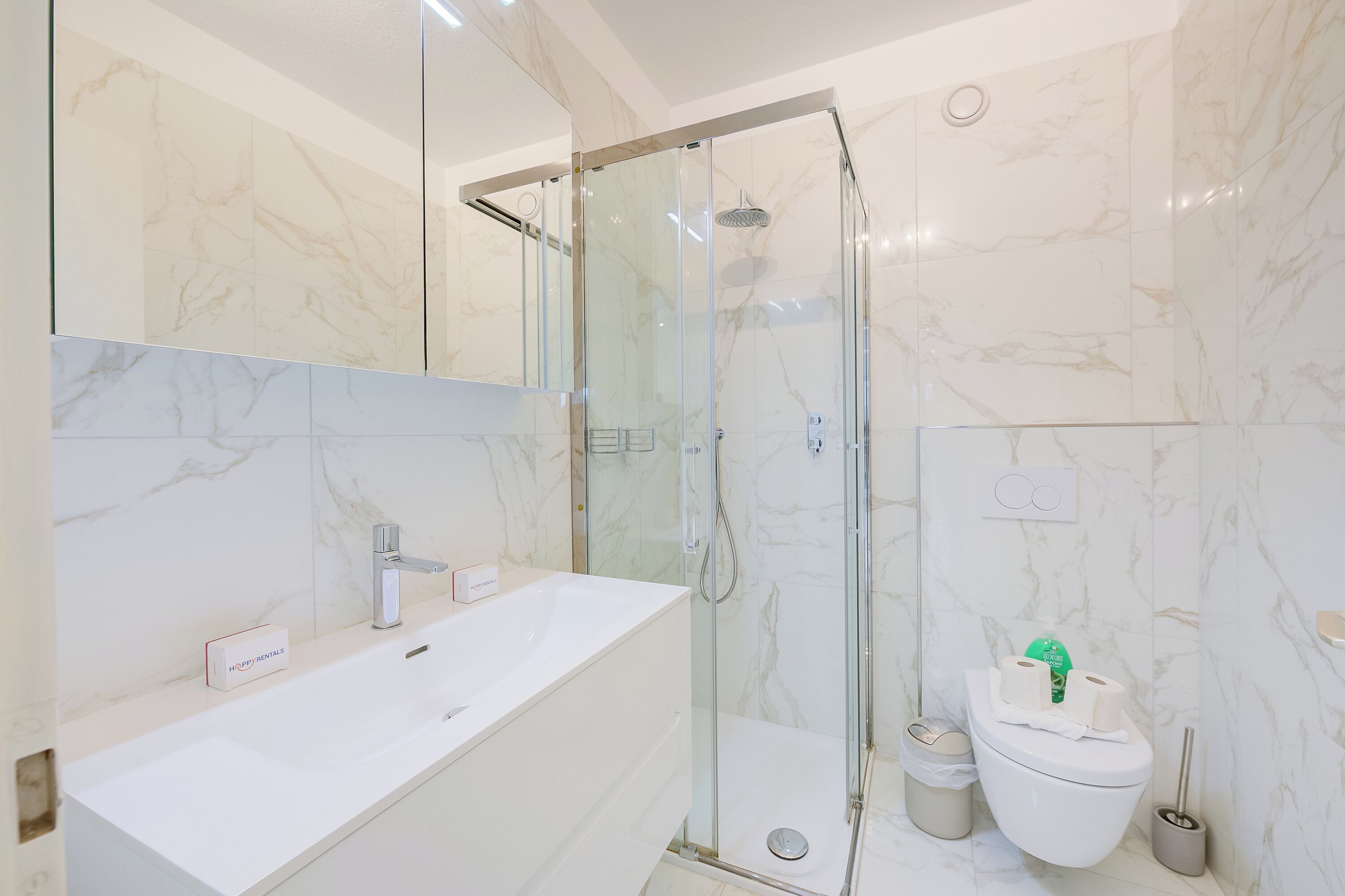 A modern bathroom with a glass-enclosed shower, a white sink, and a toilet with a green cleaning product.