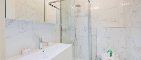A modern bathroom with a glass-enclosed shower, a white sink, and a toilet with a green cleaning product.