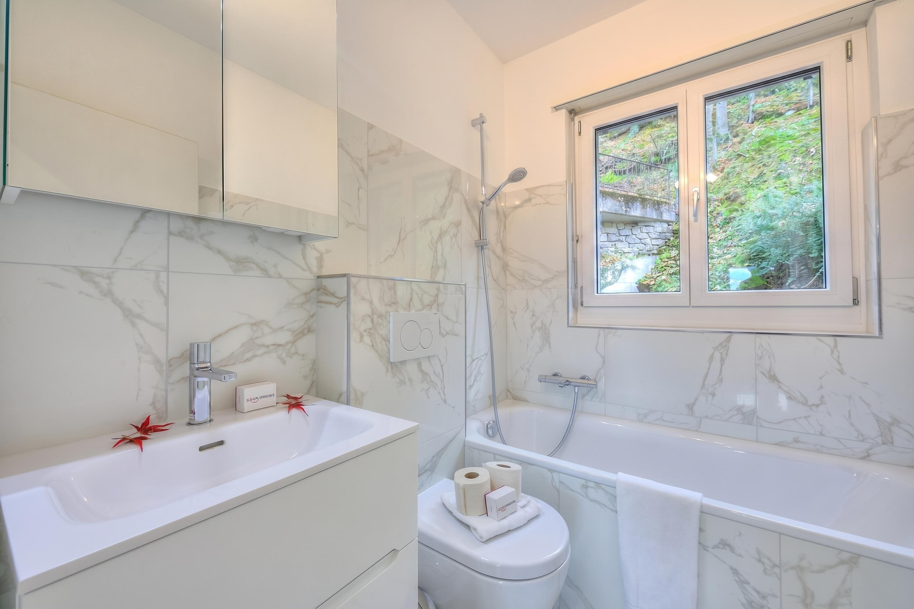 A bathroom with a marble sink, a toilet, a bathtub, and a window with a view of greenery.