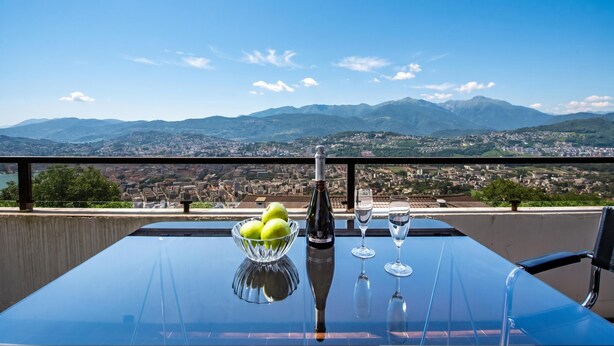 A table with a bottle, glasses, and a bowl of apples on a balcony overlooking a city and mountains. - Casa Al Bosco With Pool (Lugano)