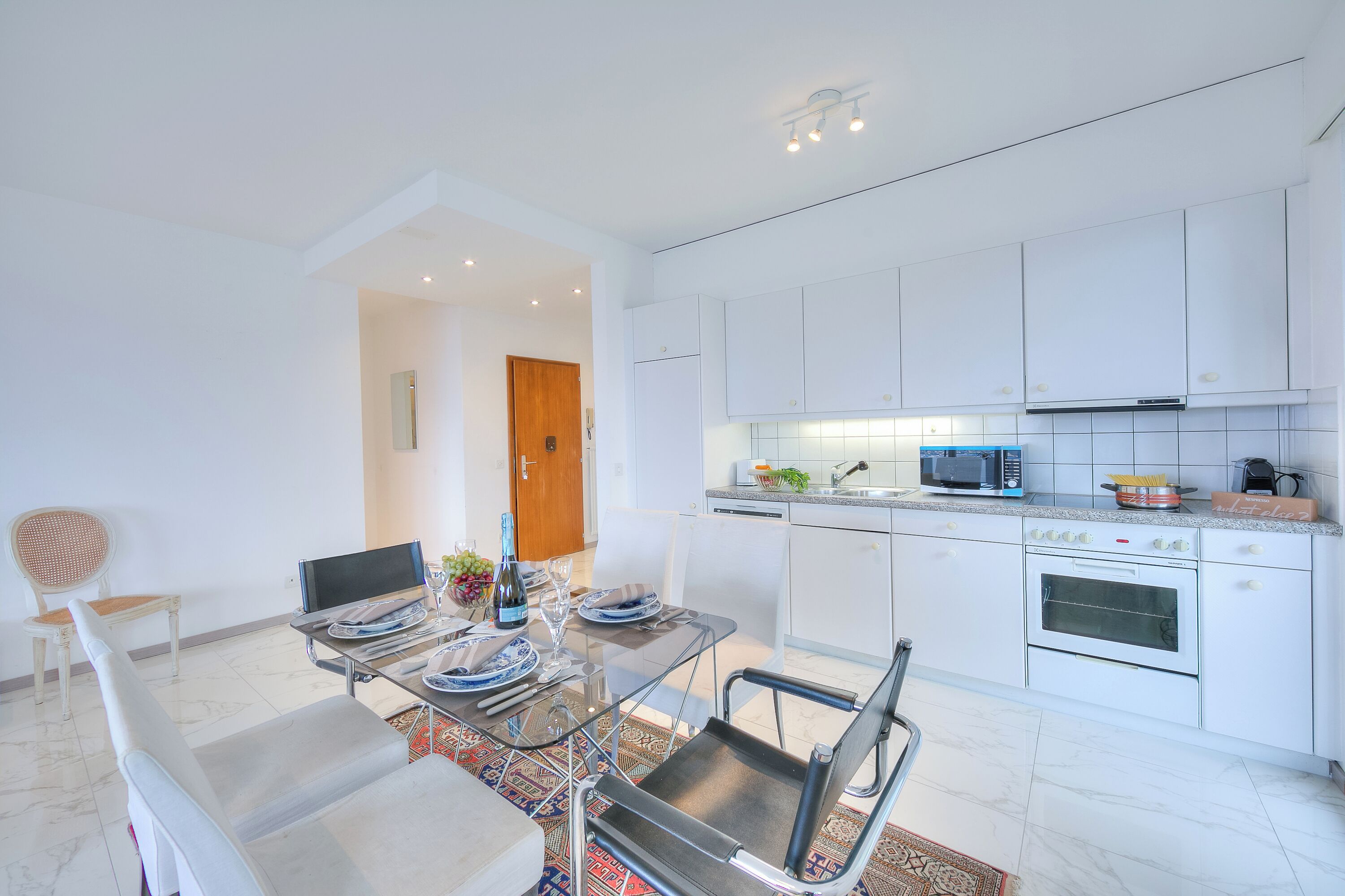 A modern kitchen with white cabinets, a built-in oven, and a microwave. There is a dining area with a glass table and chairs, and a small rug on the floor.