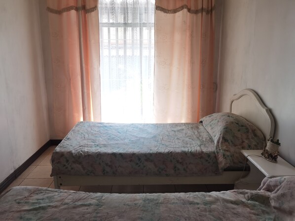 Une petite chambre avec un lit simple, une couvre-lit à motifs floraux, une table de chevet avec une lampe et une fenêtre ornée de rideaux légers.