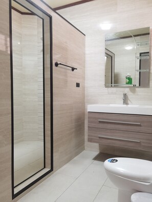 Un baño moderno con ducha de vidrio, inodoro blanco, tocador con espejo y dispensador de jabón.
