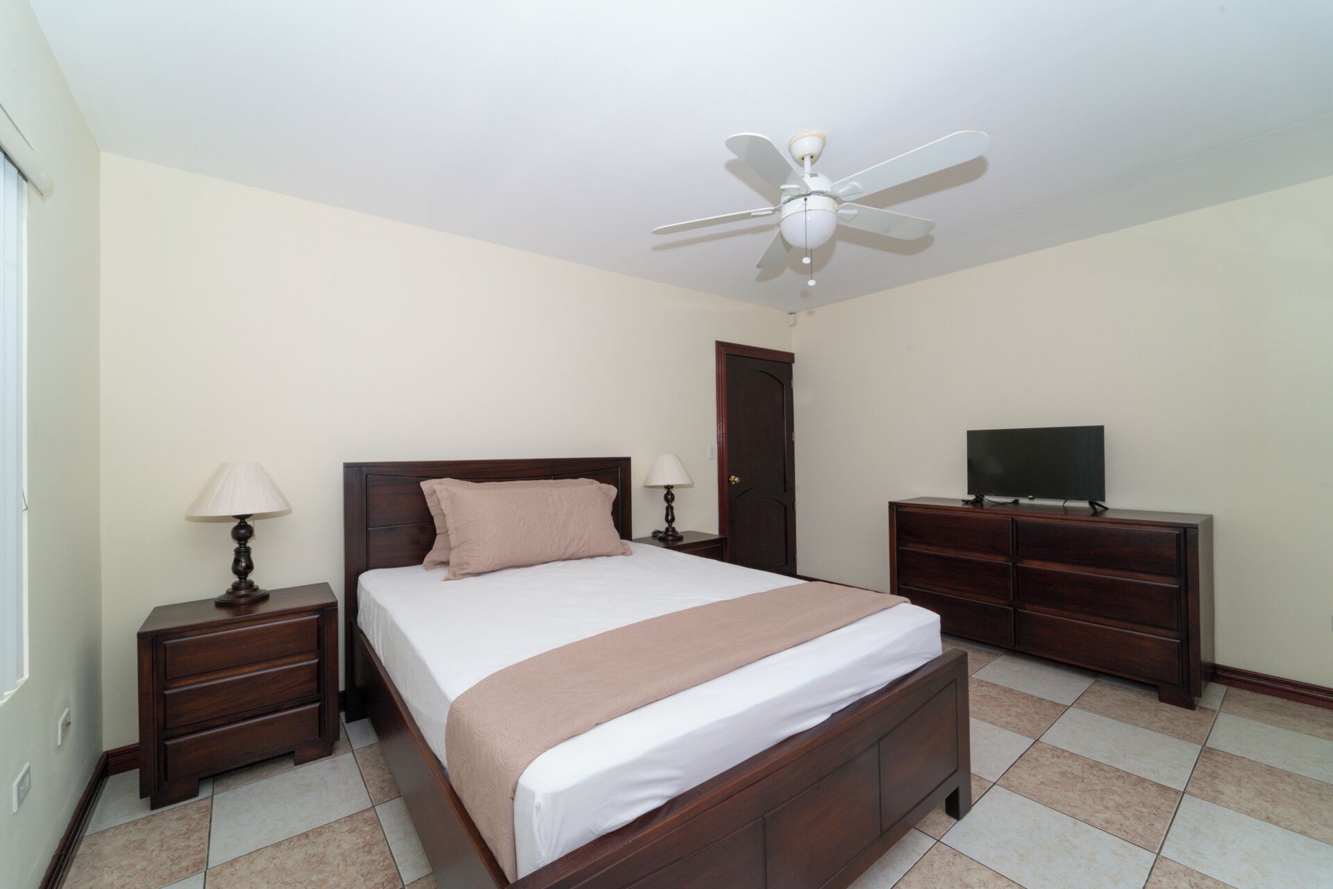 A bedroom with a bed, a ceiling fan, a TV on a dresser, and a door.