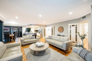 A spacious living room with a dining area, a kitchen in the background, and a large clock on the wall.